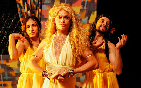 O ator Silvero Pereira como uma fada madrinha no filme Nós Somos o Amanhã. ele está com as mãos entrelaçadas no meio de duas dançarinas drag queens