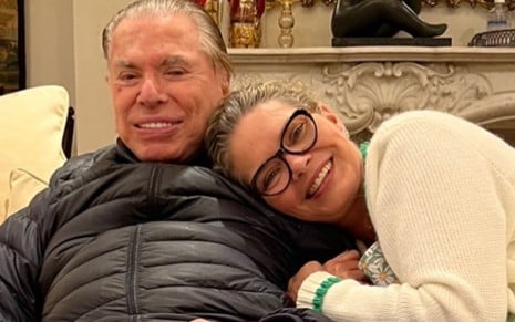 Silvio Santos e Cintia Abravanel sentados em sofá enquanto ela o abraça