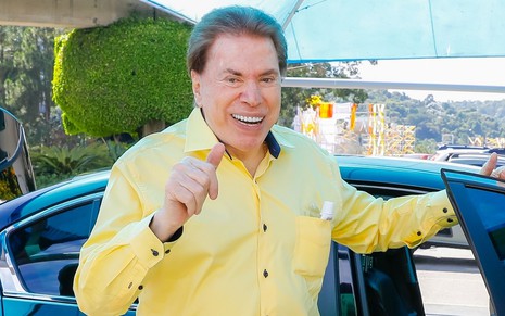 Silvio Santos de camisa amarela, em frente a um carro, sorri para foto no estacionamento do SBT, na terça-feira (26)