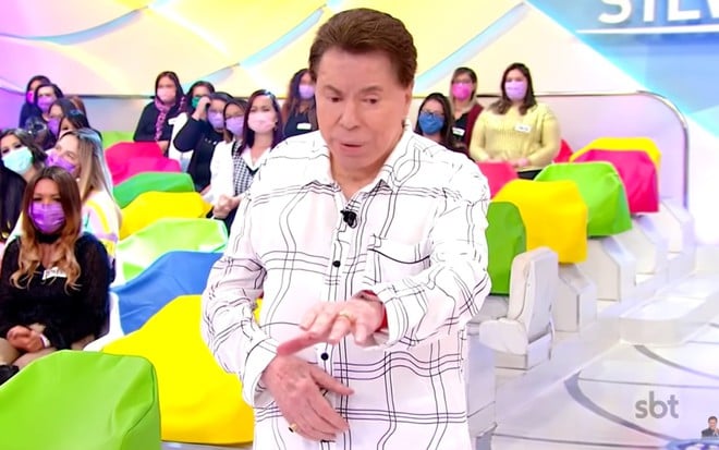 Silvio Santos de pijama branco com listras escuras, com um braço esticado, em um cenário de programa de televisão