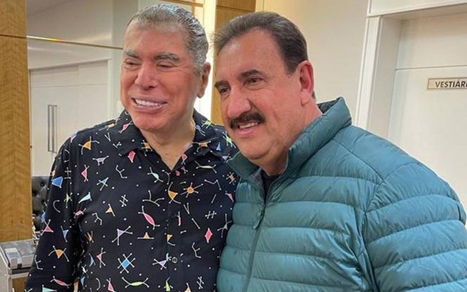 Foto de Silvio Santos e Ratinho; Silvio Santos veste camiseta preta com detalhes coloridos; Ratinho veste jaqueta azul