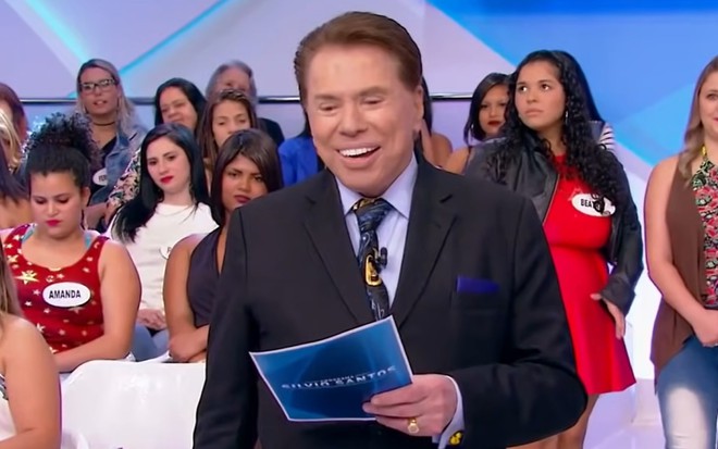 Silvio Santos sorrindo, segurando fichas de seu programa, usando um terno escuro e camisa e gravata azuis