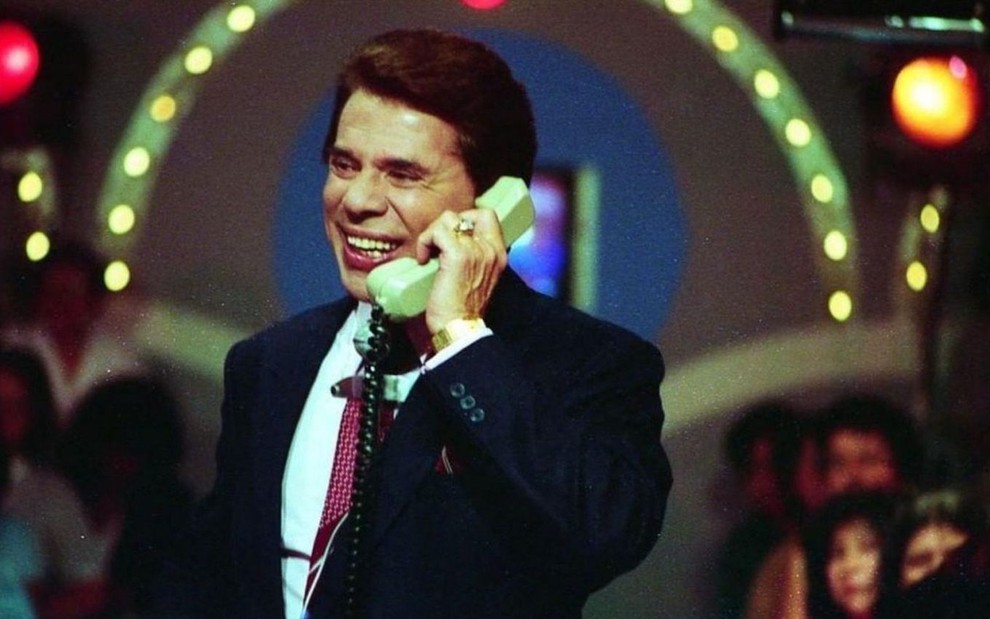 Silvio Santos fala em um telefone antigo no cenário de um programa da década de 1980