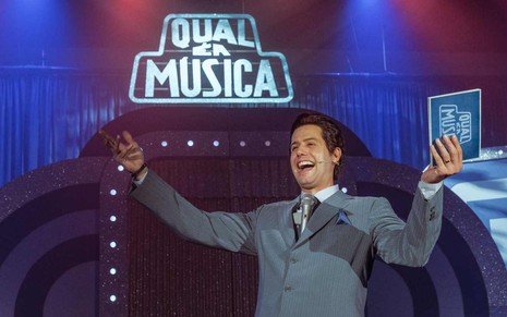 Velson D'Souza está caracterizado como Silvio Santos, com os braços abertos, à frente de uma placa do Qual É a Música?