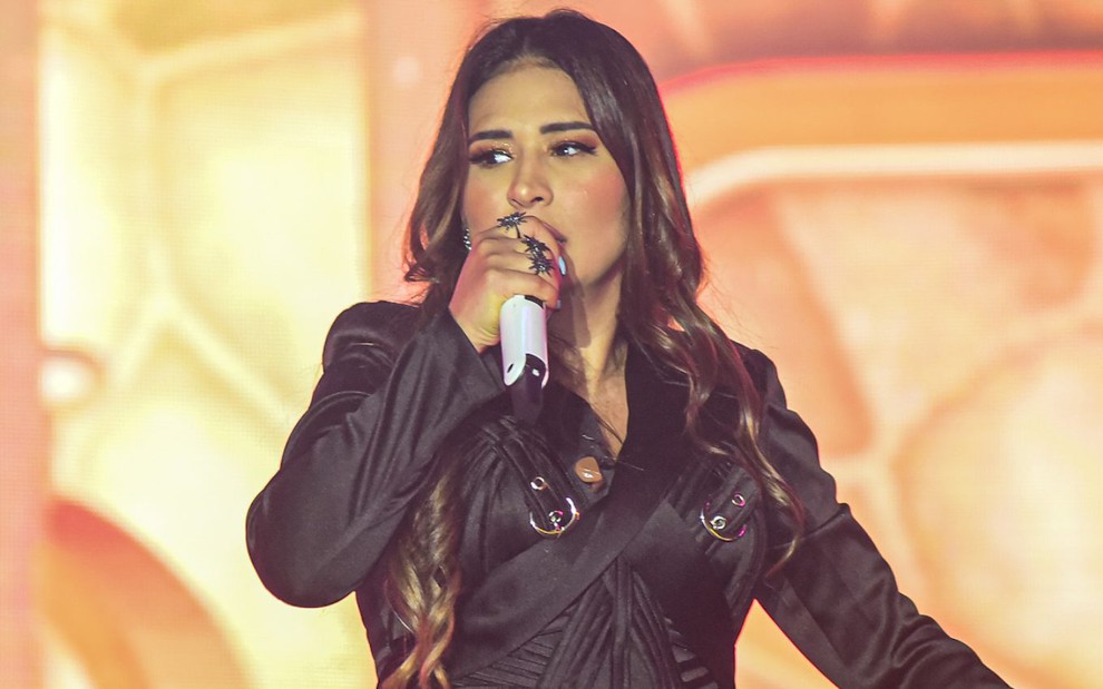 A cantora Simone Mendes durante show, com microfone branco na mão, jaqueta preta, background laranja