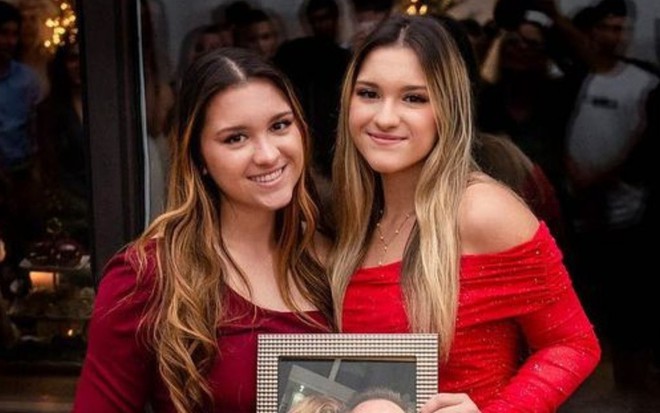 Sofia Liberato, á esquerda, e Marina Liberato, à direita. Ambas estão posando para o Instagram e vestem vestidos vermelhos. No centro, seguram um porta retrato.
