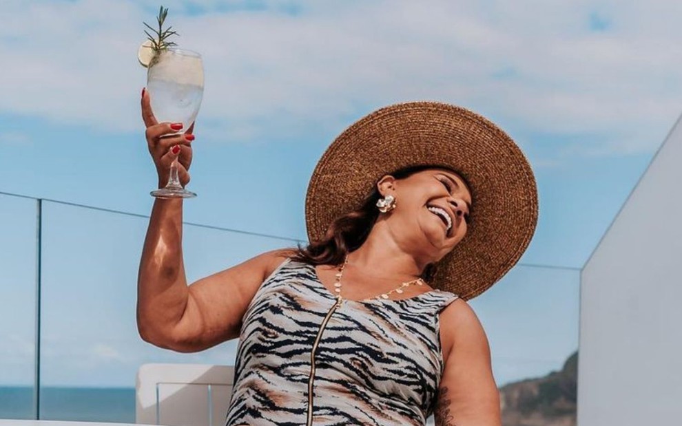 Solange Couto posa sorridente em uma paisagem de praia, com chapéu de palha e drink na mão