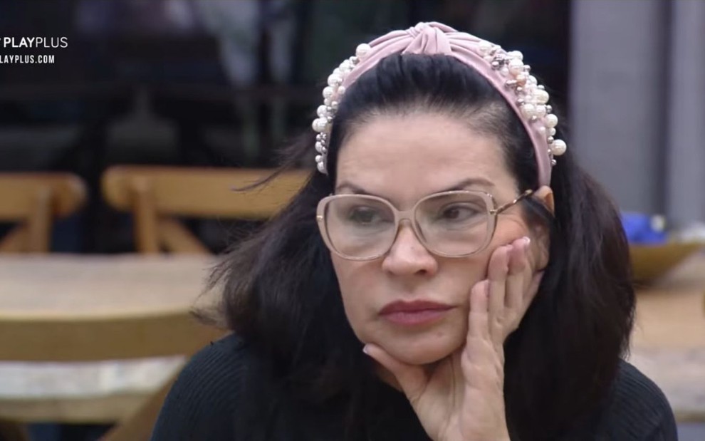 Solange Gomes na cozinha da sede de A Fazenda 13 com uma tiara rosa, camisa preta, óculos beje e um olhar de decepção e raiva