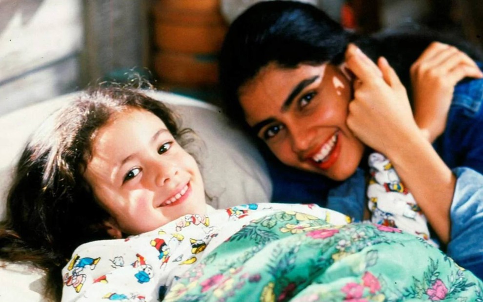 As atrizes Carolina Pavanelli e Patrícia França deitadas numa cama, sorrindo para a câmera, em set de gravações de Sonho Meu