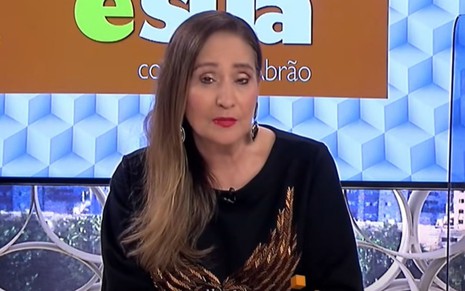 Sonia Abrão veste uma blusa preta com detalhes dourados e está com expressão séria. No fundo é possível ver o telão do programa escrito A Tarde é Sua