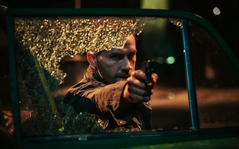 Scott Adkins segura uma arma através de um vidro de carro quebrado