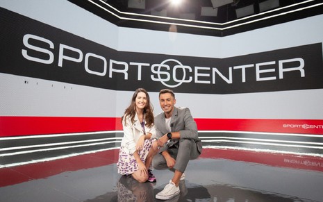Mariana Spinelli e Bruno Vicari agachados na ESPN: Mariana usa um jaleco branco com um vestido florido e tênis, enquanto Bruno usa um terno esportivo cinza e uma camisa branca