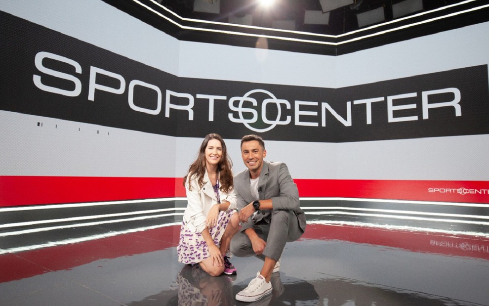 Mariana Spinelli e Bruno Vicari agachados na ESPN: Mariana usa um jaleco branco com um vestido florido e tênis, enquanto Bruno usa um terno esportivo cinza e uma camisa branca