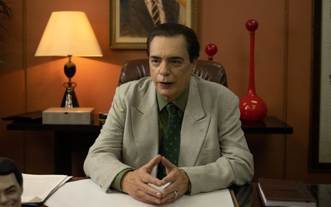 De terno e gravata, José Rubens Chachá está sentado em uma mesa e caracterizado como Silvio Santos, em cena da série O Rei da TV