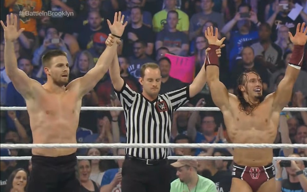 Stephen Amell e Pac tendo os braços levantados pelo árbitro na WWE