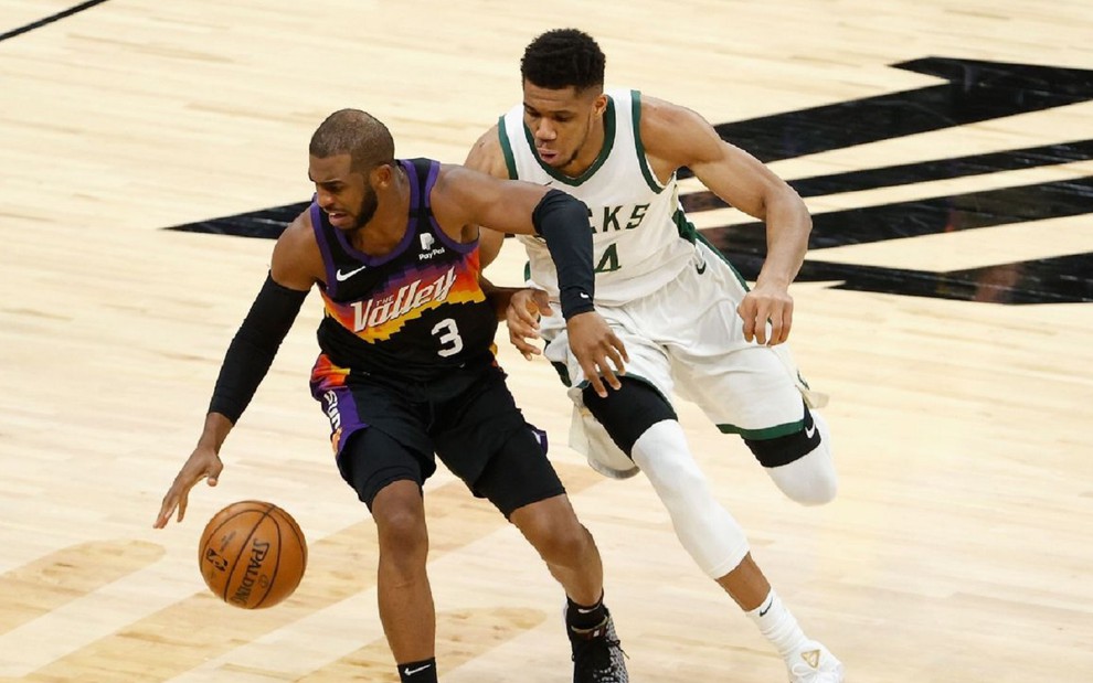 Jogador do Phoenix Suns com a bola laranja do Basquete, vestindo um uniforme todo preto. Ele é marcado por um jogador do Milwaukee Bucks, que veste branco.