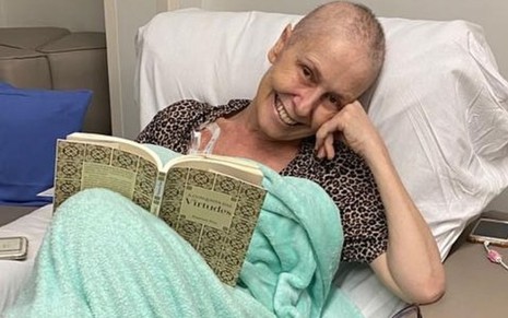 Susana Naspolini tem os cabelos raspados e usa uma blusa mais larguinha, além do lençol cirúrgico do hospital; ela está lendo um livro