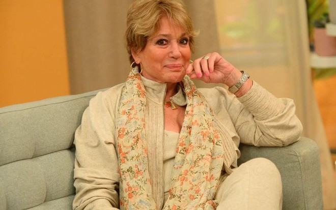 Susana Vieira sentada em sofá, com mão levemente no queixo, sorriso de boca fechada, olha para câmera
