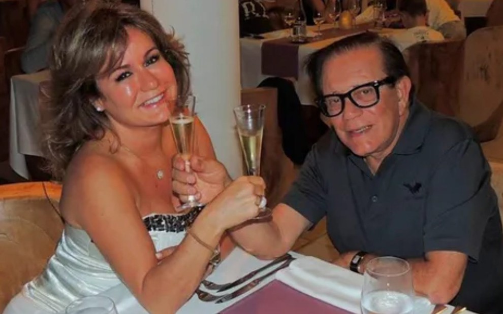 Suzy Camacho e Farid Curi brindam, com duas taças de champanhe, na mesa de um restaurante