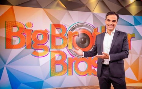 Tadeu Schmidt no estúdio do Fantástico, com uma camisa branca e ao lado do logotipo do Big Brother Brasil