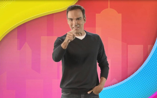 Tadeu Schmidt olha para frente e aponta para a câmera diante de fundo colorido em chamada do BBB22