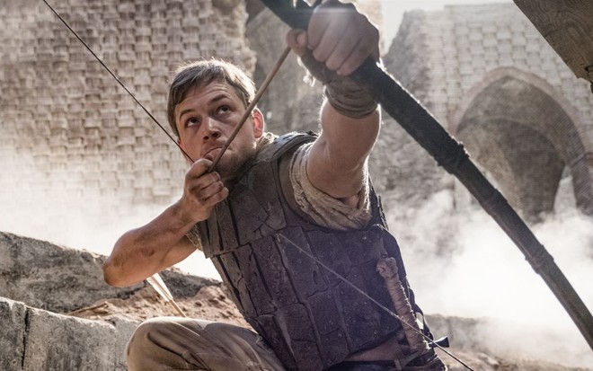 Taron Egerton agachado, segurando um arco e flecha para cima