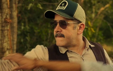 O ator Murilo Benício como Tenório em Pantanal; ele está de boné e óculos de sol olhando fixamente enquanto atira na sucuri