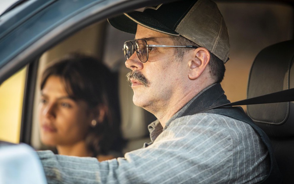 A atriz Julia Dalavia e o ator Murilo Benício como Guta e Tenório em Pantanal; eles estão dentro do carro, olhando para a  frente, com o ator no volante, de óculos de sol e boné