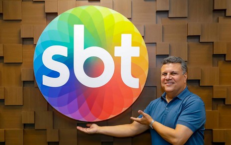Téo José, ao lado do logotipo do SBT, com uma camisa azul e apontando para a logomarca da emissora: narrador é o principal nome esportivo da emissora de Silvio Santos