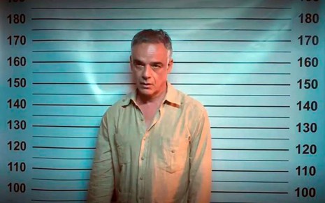 Ângelo Antônio caracterizado como Andrade; ele está na frente de um backdrop da polícia e parece confuso em cena de Terra e Paixão