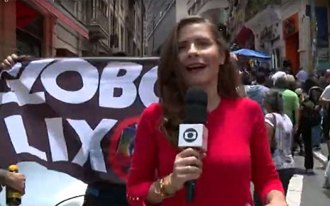 Thaís Luquesi durante reportagem para o SP1; atrás está um cartaz "Globo Lixo"