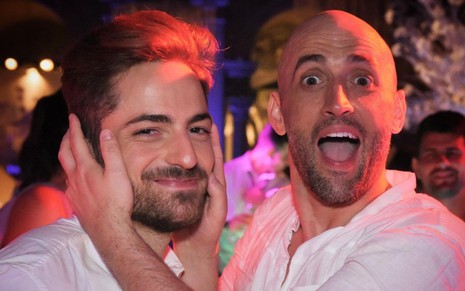 Thales Bretas (à esquerda) sorri discretamente; Paulo Gustavo (à direita) com expressão surpresa e mãos no rosto do marido