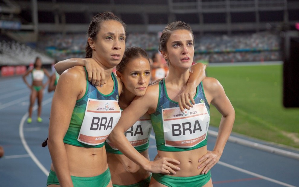 Thalita Carauta, Priscila Steinman e Fernanda de Freitas em uma pista de corrida, com roupas de velocistas, abraçadas, olhando para alguma coisa