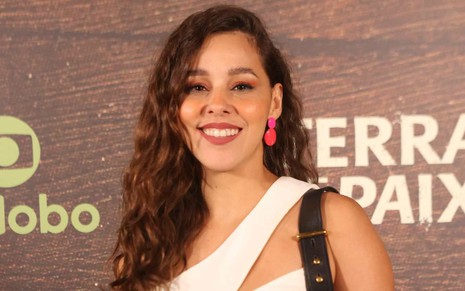 Thati Lopes sorri na festa de lançamento de Terra e Paixão e está na frente do painel com o logo da novela