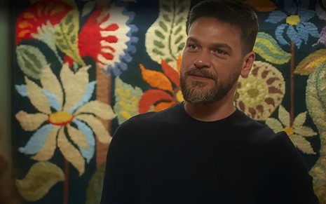 Em cena de Vai na Fé, Emilio Dantas usa blusa preta está falando com alguém em um fundo com painel de flores pintadas