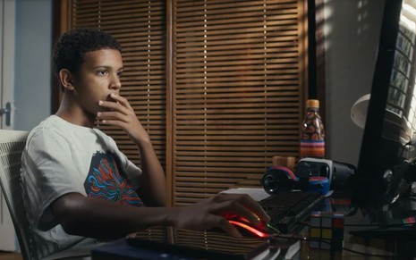 O ator Ricardo Santos como Theo em Travessia; ele está sentado de frente a um computador, enquanto olha para a tela com a mão na boca