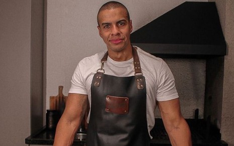 O apresentador Thiago Oliveira de avental de cozinheiro, camiseta branca, expressão séria