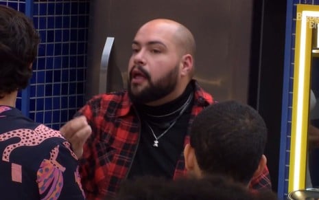 Em frente a geladeira, Tiago Abravanel discursa para colegas usando camisa xadrez vermelha e preta