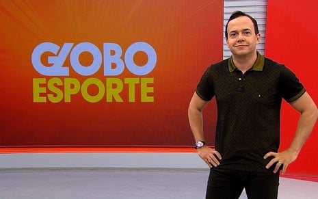 Tiago Medeiros está todo de preto, no cenário laranja do Globo Esporte no Recife (PE); ele sorri para a câmera e feliz
