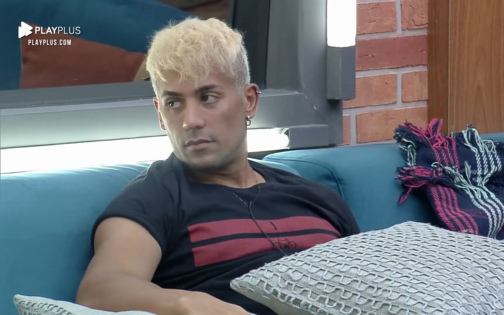 Com cabelos platinados, Tiago Piquilo faz cara de reflexivo enquanto está sentado no sofá do reality