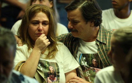 Em cena da série Todo Dia a Mesma Noite, Debora Lamm e Thelmo Fernandes estão com cara de choro, usando uma camisa com a foto da filha que morreu no incêndio da boate Kiss