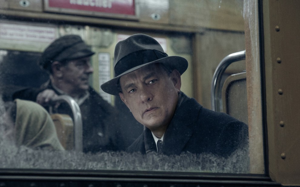 Tom Hanks com um chapéu, dentro de um trem, olhando pela janela