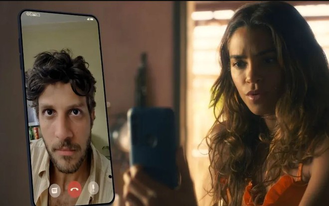 Os atores Chay Suede e Lucy Alves como Ari e Brisa em Travessia; ela está olhando para o celular enquanto conversa com ele, com cara de assustada e ele aparece do lado esquerdo da foto na tela do celular, olhando para ela