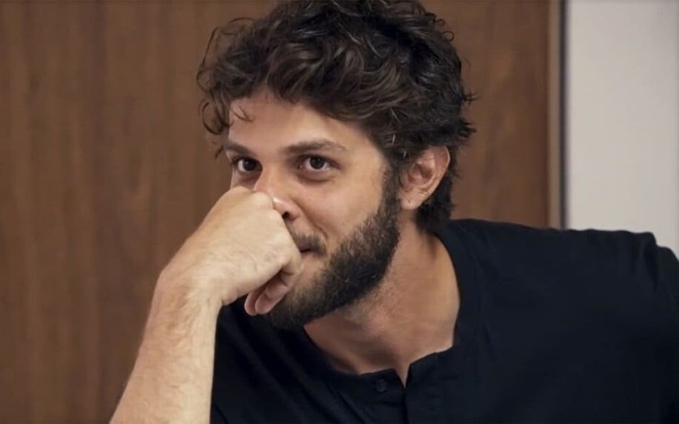 Em cena de Travessia, Chay Suede usa blusa preta, sorrindo, com a mão apoiada no nariz e na boca