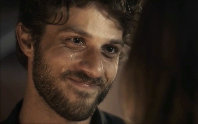Em cena de Travessia, Chay Suede usa blusa preta e está falando com alguém, sorrindo e chorando
