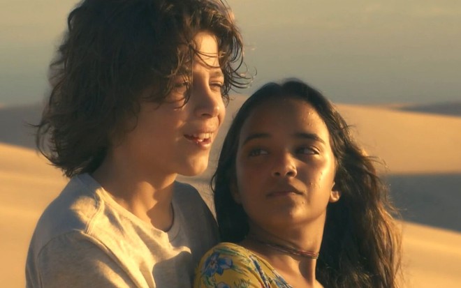 Os atores João Bravo e Mariah Yohana como Ari e Brisa em Travessia; eles estão abraçados, enquanto ele está atrás olhando para frente e ela olhando para ele