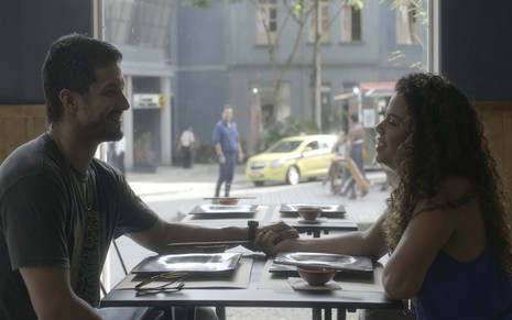 Em cena de Travessia, Romulo Estrela segura a mão de Clara Buarque, sentados, frente a frente, em um restaurante; Rodrigo Lombardi observa de fora