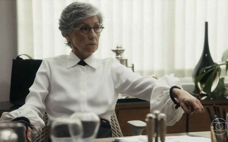 Em cena de Travessia, Cassia Kis está sentada na cadeira de sua sala; ela usa blusa branca de botão