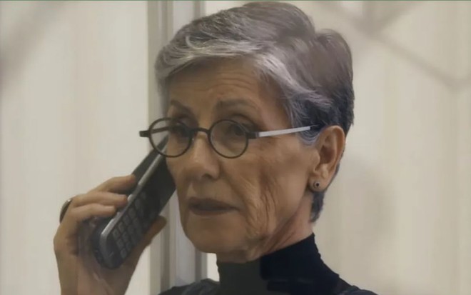 A atriz Cassia Kis como Cidália em Travessia; ela está ligeiramente virada para o lado, enquanto olha para frente com cara de espanto e segura um telefone
