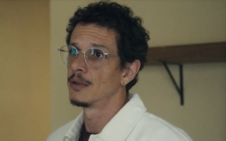 Em cena de Travessia, Rafael Losso usa casaco branco, óculos e está falando com alguém com expressão de preocupação